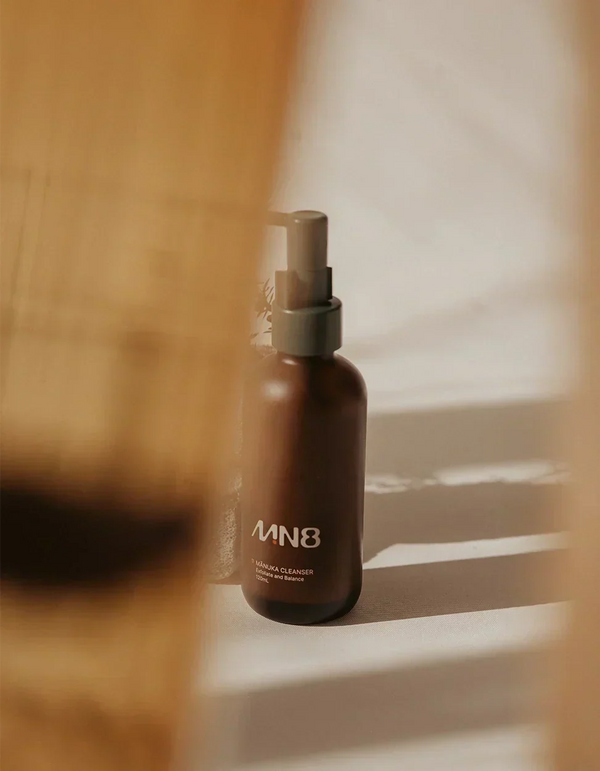 Alt text: MN8 Mānuka Exfoliate Cleanser 120ml pump bottle on warm-toned bathroom shelf
