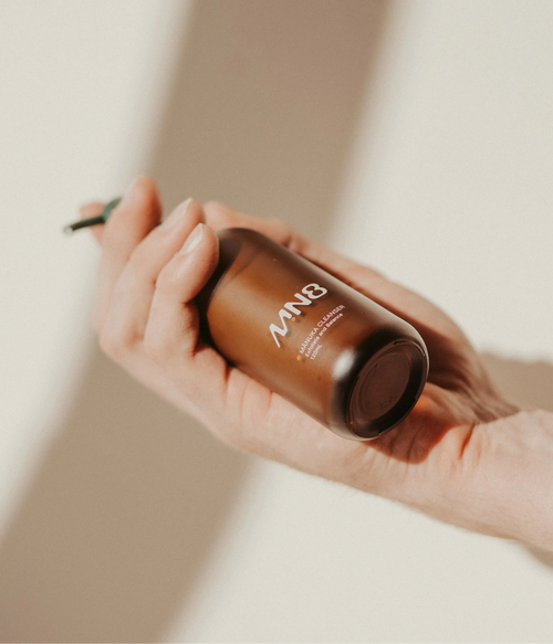 Hand holding MN8 Mānuka Exfoliate Cleanser amber pump bottle against warm neutral background
